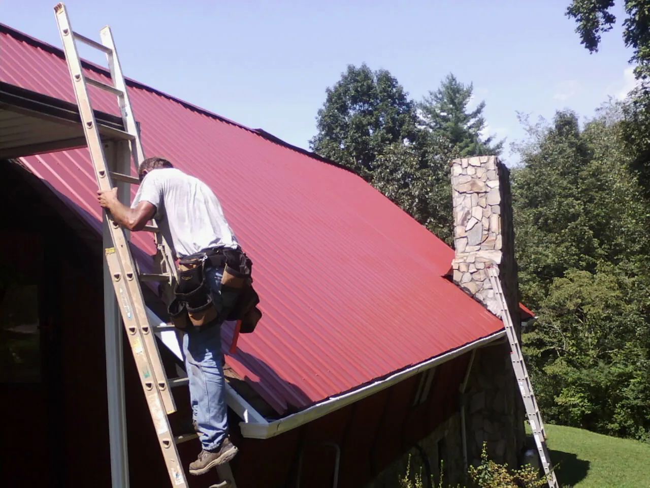 Roofer on a ladder beside a red metal roof and stone chimney — Soffit & Fascia Repair by Roofing Brothers in Lackawaxen